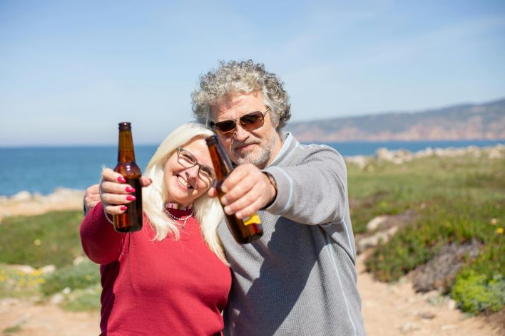 Senior couple smiling and clinking beer bottles by the coastline on a sunny day.