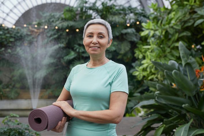 Elderly woman holding yoga mat and smiling in a lush greenhouse setting.