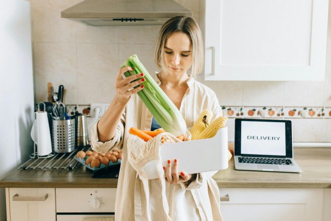 Woman unpacks fresh vegetables from delivery box in a modern kitchen setup with a laptop nearby.