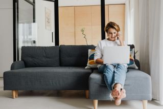 Woman sitting barefoot on a sofa, working on a laptop and talking on the phone in a modern home setting.