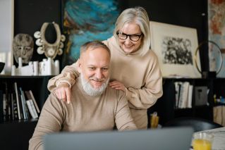 A happy elderly couple wearing sweaters, smiling while using a laptop indoors.